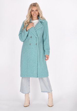 Teal, fluffy, double-breasted coat with a collar, featuring silver buttons, worn over light denim flared pants and beige ankle boots.