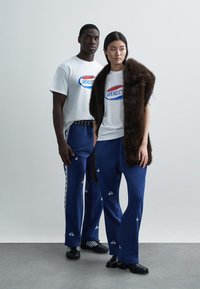 Dois modelos vestindo roupas casuais: uma T-shirt branca com um gráfico azul e vermelho, calças azuis com padrão e um colete de pelo sintético castanho.