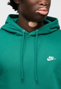 Zelena bombažna kapa s kenguru žepom, vrvicami in belim logotipom Nike na prsih. Mehka tekstura in priložnostna zasnova.