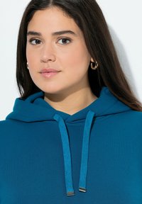 Felpa blu verde con tasca frontale a kangaroo, cordini e dettagli argento. Tessuto liscio con una vestibilità comoda e polsini a costine.