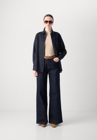 Donkerblauwe wijde jeans gecombineerd met een beige top en een marineblauwe button-up blouse. Een bruine leren riem en een zonnebril maken de look compleet.