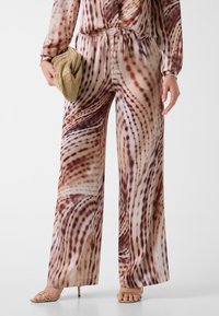 Weit geschnittene Hosen aus einem glatten, leichten Stoff mit einem wirbelnden Muster in Brauntönen und Creme. Kombiniert mit einer goldenen Clutch und Sandalen.