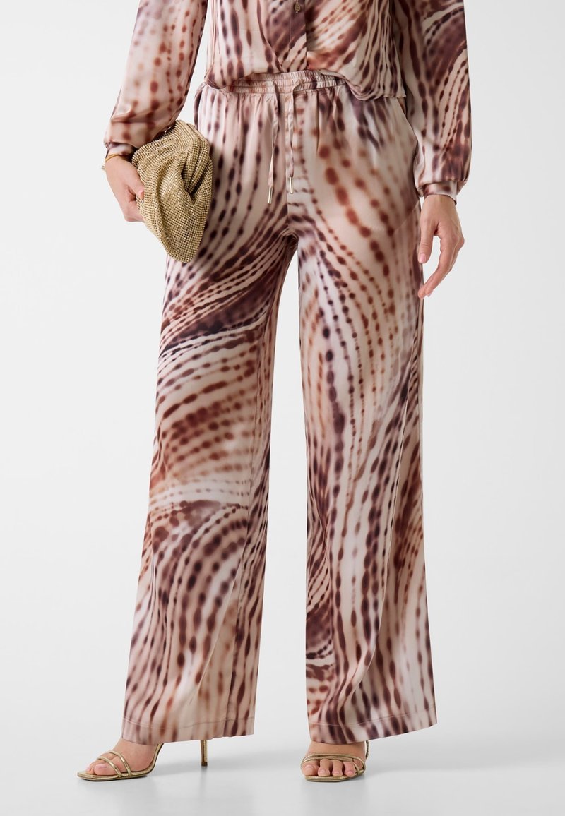 Weit geschnittene Hosen aus einem glatten, leichten Stoff mit einem wirbelnden Muster in Brauntönen und Creme. Kombiniert mit einer goldenen Clutch und Sandalen.