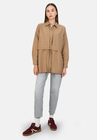 Light brown oversized shirt with a cropped design and tie detail at the waist, paired with light blue jeans and burgundy sneakers.