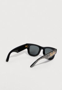 Occhiali da sole neri con lenti grigie, montatura in plastica lucida e aste decorate con scritte dorate e accenti metallici.