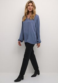 Lyseblå langærmet bluse med V-hals og tekstureret stof, parret med sorte slim-fit bukser og sorte hæl-støvletter.
