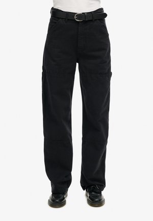 Zwarte loszittende broek met steekzakken aan de voorkant en een zwarte riem, gecombineerd met zwarte schoenen bij een persoon die een wit shirt draagt.