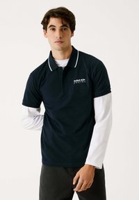 Marine polo-shirt med hvid krave og kant, prydet med et Red Bull Racing-logo. Båret over en hvid langærmet skjorte, lavet af letvægtsstof.