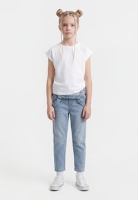 Fată cu păr blond prins în cocuri duble și împletituri, purtând un tricou alb, blugi bleu deschis, șosete albe și adidași albi, stând în picioare.