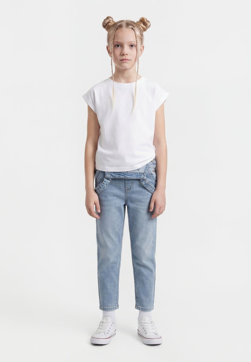 Fată cu păr blond prins în cocuri duble și împletituri, purtând un tricou alb, blugi bleu deschis, șosete albe și adidași albi, stând în picioare.