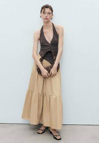 Top a fascia marrone, abbinato a una gonna maxi beige a strati. Tiene in mano una borsa marrone scuro con dettagli argentati. Sandali neri con cinturini completano l'outfit.