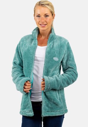 Femme blonde portant une veste zippée duveteuse bleu sarcelle sur une chemise blanche, tenant la veste ouverte, debout devant un fond blanc.
