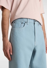 Light blue denim jeans with a straight cut, featuring visible stitching, five pockets, and black metal button accents. Cotton fabric texture.