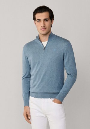 Joven con suéter azul claro de media cremallera sobre una camisa blanca y pantalones blancos, de pie con una mano en el bolsillo.