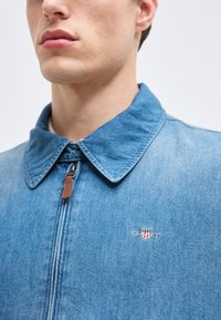 Άνδρας που φοράει ανοιχτό μπλε τζιν μπουφάν με γιακά και ημιδιάφανο φερμουάρ, με ένα μικρό κεντητό κόκκινο και λευκό σήμα ασπίδας στο στήθος.