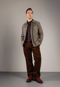 Šedá semišová bunda se dvěma předními kapsami, tmavý svetr, hnědé kordové kalhoty a červené šněrovací boty s černými podrážkami. Neutrální pozadí.