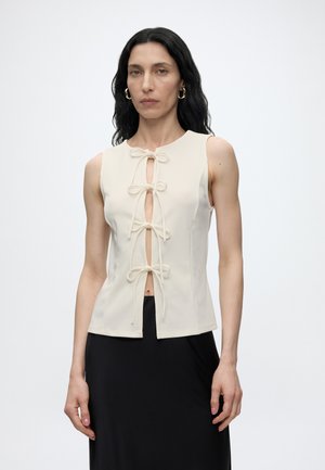 Femme aux longs cheveux foncés portant un haut beige sans manches avec quatre nœuds devant et une jupe noire, debout devant un fond clair uni.