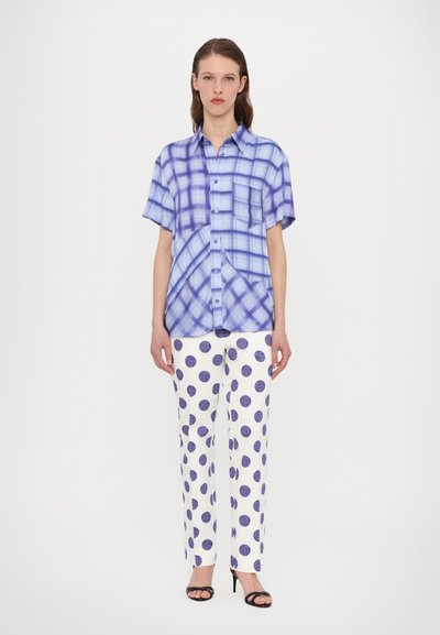 Camicia a maniche corte con bottone a fantasia a scacchi blu, abbinata a pantaloni bianchi con grandi pois viola e tacchi aperti neri.