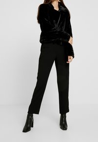 Woman wearing black faux fur zip-up jacket, black straight-leg pants, and black heeled ankle boots, standing on a plain white background.