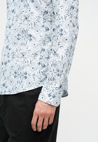 Personne portant une chemise blanche à manches longues avec un motif floral bleu et un pantalon noir, la main détendue le long du corps.