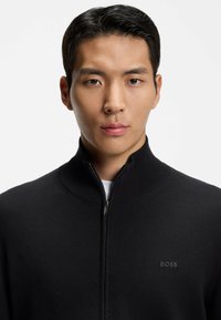 Młody mężczyzna o krótkich czarnych włosach, ubrany w czarny sweter zapinany na zamek z logo BOSS na piersi, stoi przodem na białym tle.