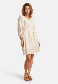 Helles beiges Kleid mit einem Zickzackmuster, V-Ausschnitt und Dreiviertelärmeln; verfügt über einen passenden Taillengürtel und eine weiche Stoffstruktur.