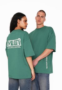 T-shirt verdi oversize con grande logo bianco sul retro e accenti testuali, realizzata in tessuto morbido, abbinata a jeans in denim blu.