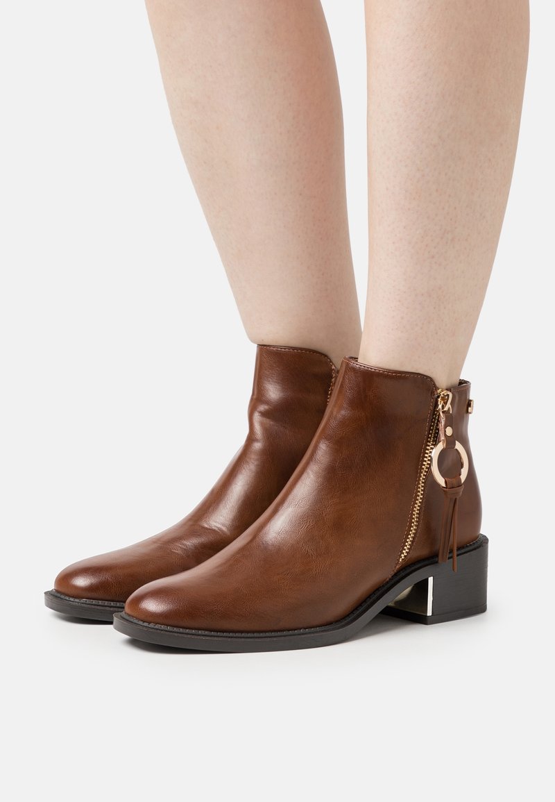 Bottes courtes en cuir marron avec fermeture éclair latérale et breloque anneau doré décorative, portées sur des jambes nues avec des talons blocs bas.