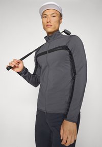 Gray zip-up golf jacket with black side stripes and logo; features a high collar and fitted design. Model holds a golf club, wearing a white cap.
