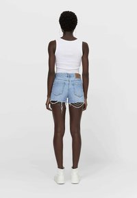 Shorts vaqueros, azul claro con dobladillo deshilachado, diseño de cinco bolsillos, parche trasero de cuero marrón. Combinados con una camiseta sin mangas blanca y zapatos blancos.