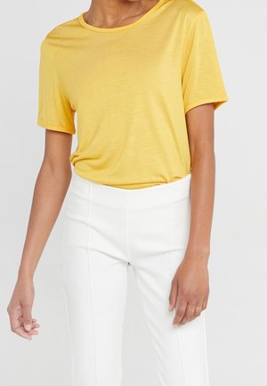Donna che indossa una maglietta gialla a maniche corte infilata in pantaloni bianchi a vita alta, in piedi davanti a uno sfondo chiaro e uniforme.