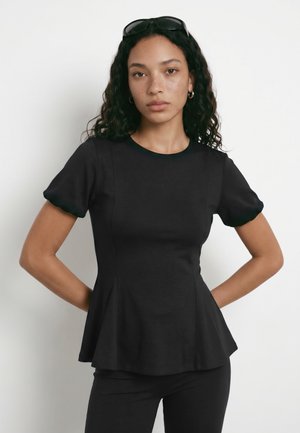 Femme aux cheveux bouclés portant un top péplum noir ajusté et un pantalon noir, lunettes de soleil posées sur la tête, debout les mains sur les hanches.