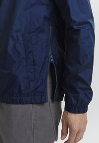 Veste légère bleu marine avec une poche latérale zippée, des poignets élastiques et un ourlet élastique. Le tissu a une texture lisse et brillante.