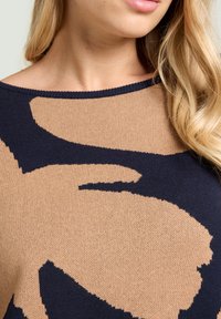 Maglione lavorato a maglia con un design astratto blu navy e beige, dotato di un ampio scollo e una texture morbida. Il motivo crea un forte contrasto visivo.