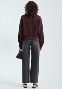 Vrouw met krullend haar, een wijnrode kabeltrui, zwarte wijde jeans, die een kleine zwarte handtas draagt en zwarte sandalen aan heeft, met haar rug naar de camera toegekeerd.