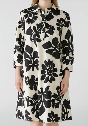 Mujer que lleva un vestido de largo hasta la rodilla, con mangas largas, grandes estampados florales negros sobre fondo blanco y un cuello con botones.