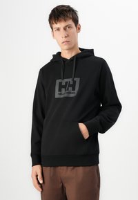 Schwarzer Kapuzenpullover mit einer vorderen Taschenklappe, Kordelzugkapuze und einem grauen Logo, das "HELLY HANSEN" liest. Hergestellt aus weichem, glattem Stoff.
