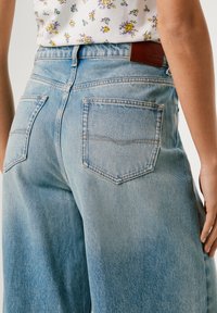 Svijetloplavi traperice s visokim strukom, detaljnim stražnjim džepovima i smešnim kožnim oznakom branda. Imaju opušteni kroj i izblijedjeli izgled.