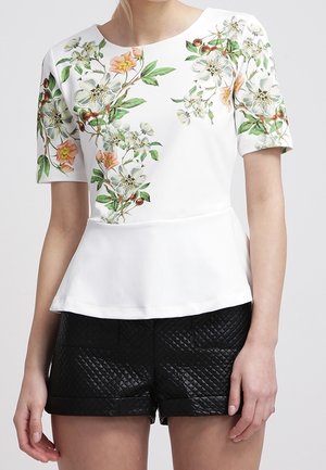 Weiße kurzärmlige Bluse mit buntem Blumenmuster kombiniert mit schwarzen gesteppte Shorts an einer stehenden Person.