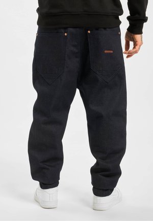 Personne portant un pantalon en denim noir ample avec de grandes poches arrière et des baskets blanches, debout devant un fond uni.