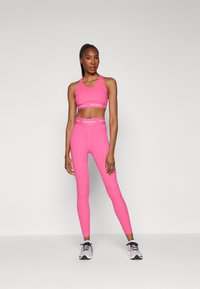 Ružičasti atletski crop top i tajice izrađeni od rastezljivog materijala s vidljivim brendingom. Imaju uski dizajn i glatku teksturu.