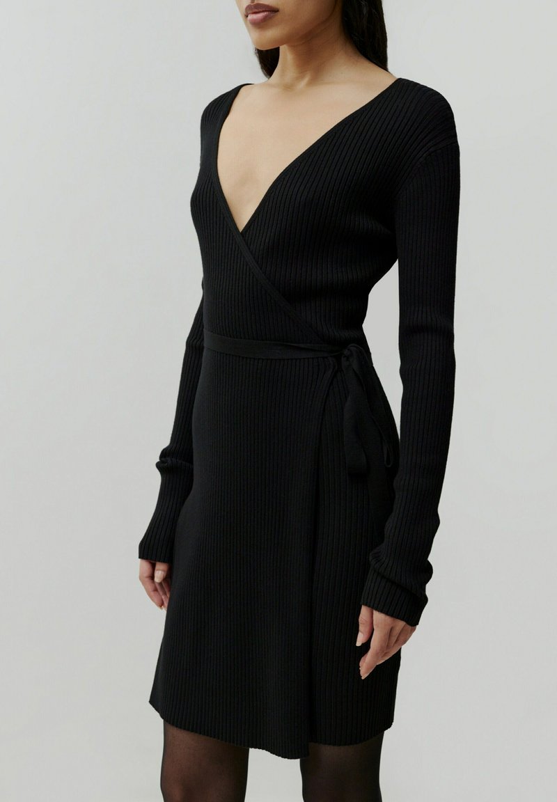 Schwarzes geripptes Wickelkleid mit tiefer V-Ausschnitt und langen Ärmeln. Hat eine Krawatte in der Taille und eine figurbetonte Silhouette.