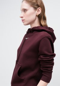 Burgundska kapuca s prednjim žepom i podesivim vezicom na kapuljači. Sadrži mali bijeli Nike logo na prsima. Mekani materijal i opušteni kroj.
