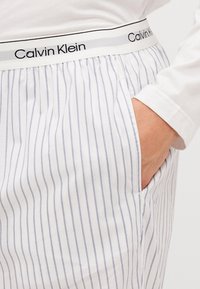 Baltas strīpains pidžamu bikses ar gaiši ziliem vertikāliem svītrainiem, kas aprīkotas ar elastīgu jostasvietu ar "Calvin Klein" logo un sānu kabatām.