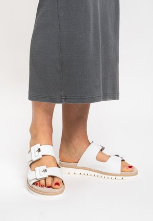 Nohy v bílých sandálech s dvojitými pásky a přezkami, s červeným lakem na nehty, v kombinaci se šedými kotníkovými kalhotami na bílém pozadí.