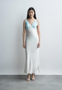 Žena s dlhými tmavými vlasmi, oblečená v bielych saténových nočných šatách s svetlomodrými čipkovými detailmi, stojaca pred jednoduchou bielou stenou.