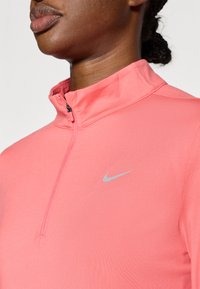 Nike zip-up top in felroze stof met een hoge kraag, voorzien van een zilveren Nike-logo op de linkerborst, gladde textuur.