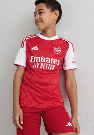 Červený futbalový dres s krátkym rukávom a bielymi akcentmi, obsahujúci logo Arsenalu a text "Emirates Fly Better", v kombinácii s rovnakými červenými šortkami.