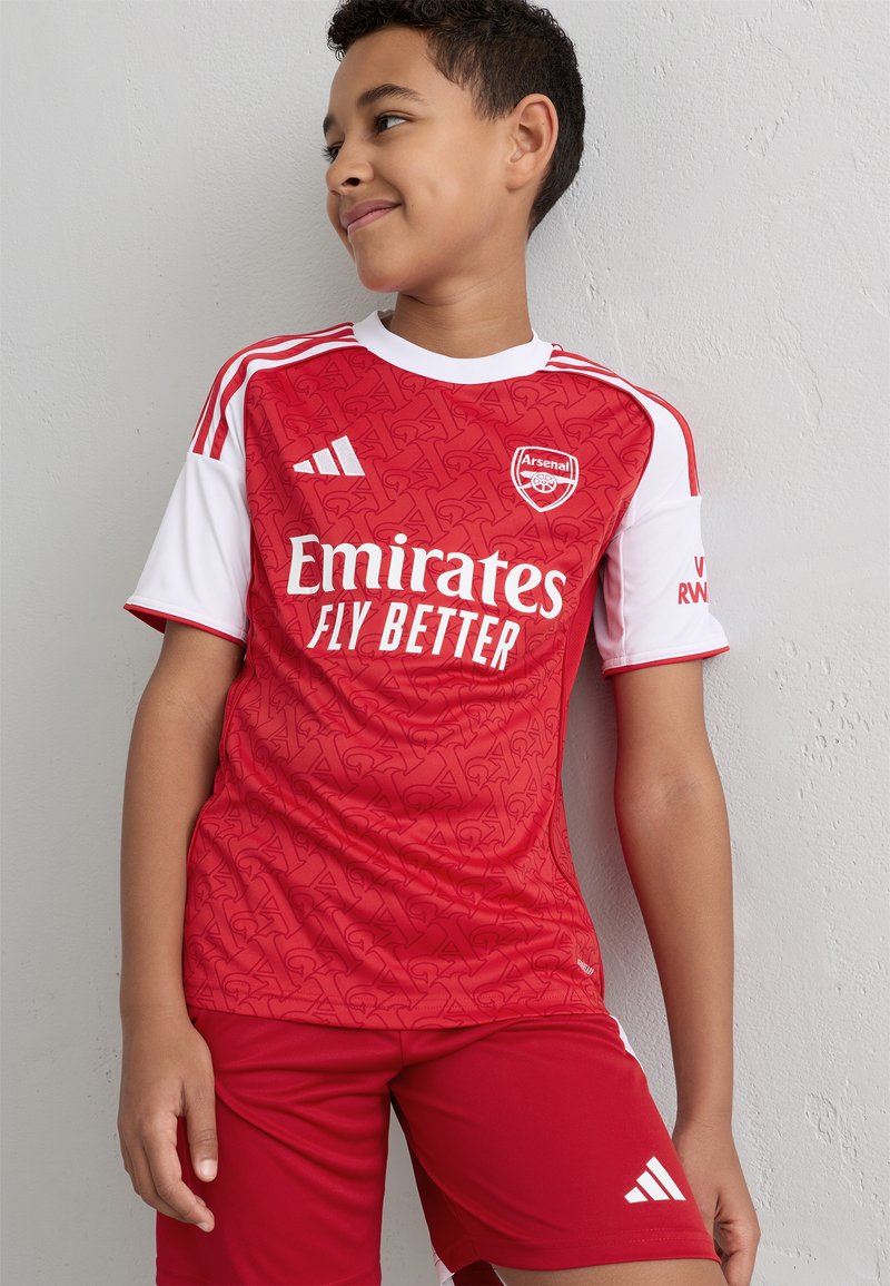 Rød fotballtrøye med kort erme og hvite detaljer, med Arsenal-logoen og teksten "Emirates Fly Better", kombinert med matchende røde shorts.