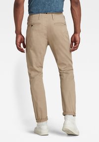 Homme portant un pantalon chino beige avec les revers roulés, des baskets blanches et une chemise bleue à manches courtes, vu de dos sur fond blanc.